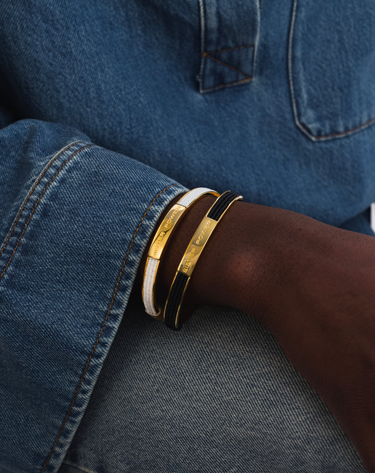 A close-up shot of a model's wrist, styled with two gold Sister Power bracelets stacked together, one with white beads and one with black beads.