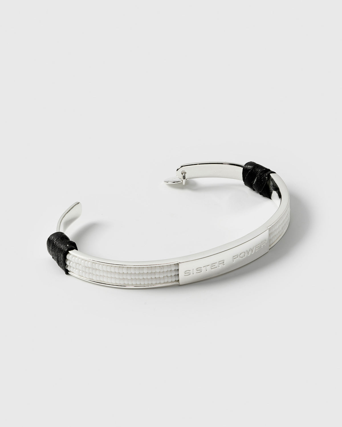 A product shot of the Sister Power Bracelet Silver. The silver cuff is adorned with rows of small white beads and features a central plate engraved with the words 'SISTER POWER'.