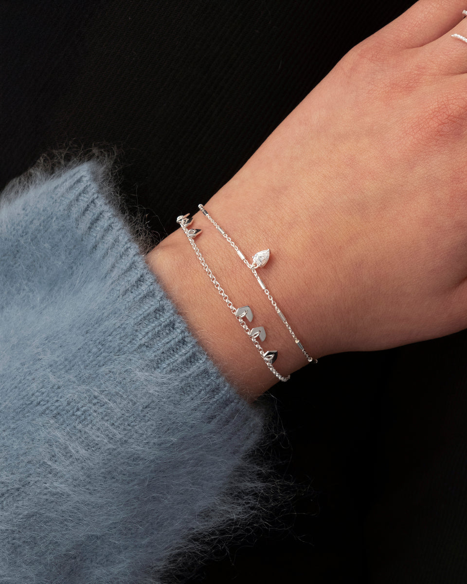 A model's wrist adorned with two stacked silver bracelets from Syster P. The Layers Bianca Bracelet features small, grouped leaf-like charms, and it is styled with another delicate chain bracelet that has a single crystal charm.