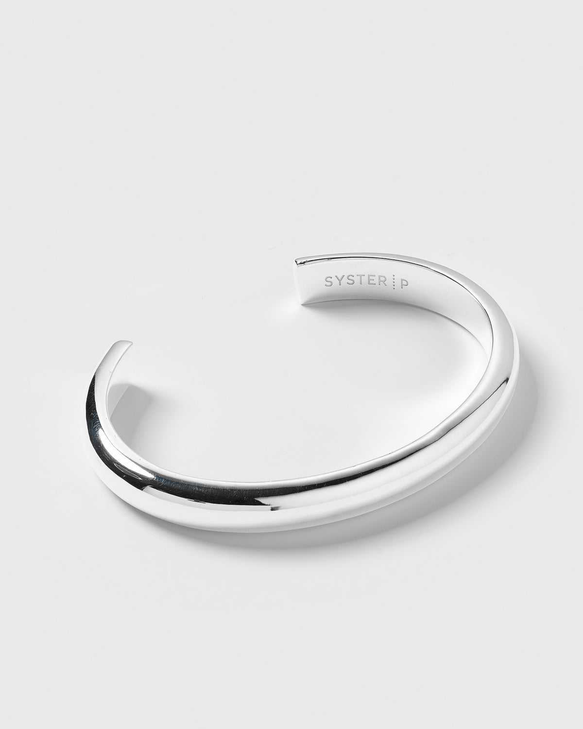 A product shot of the Bolded Bangle in silver, a chunky and smooth cuff-style bracelet with the Syster P logo engraved on the inside.