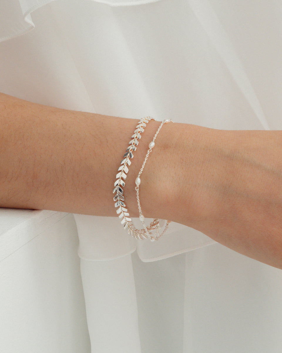 A model wears the Treasure Multi Pearl Bracelet in silver, a delicate chain with small freshwater pearls, styled with another silver bracelet featuring a repeating chevron link design.