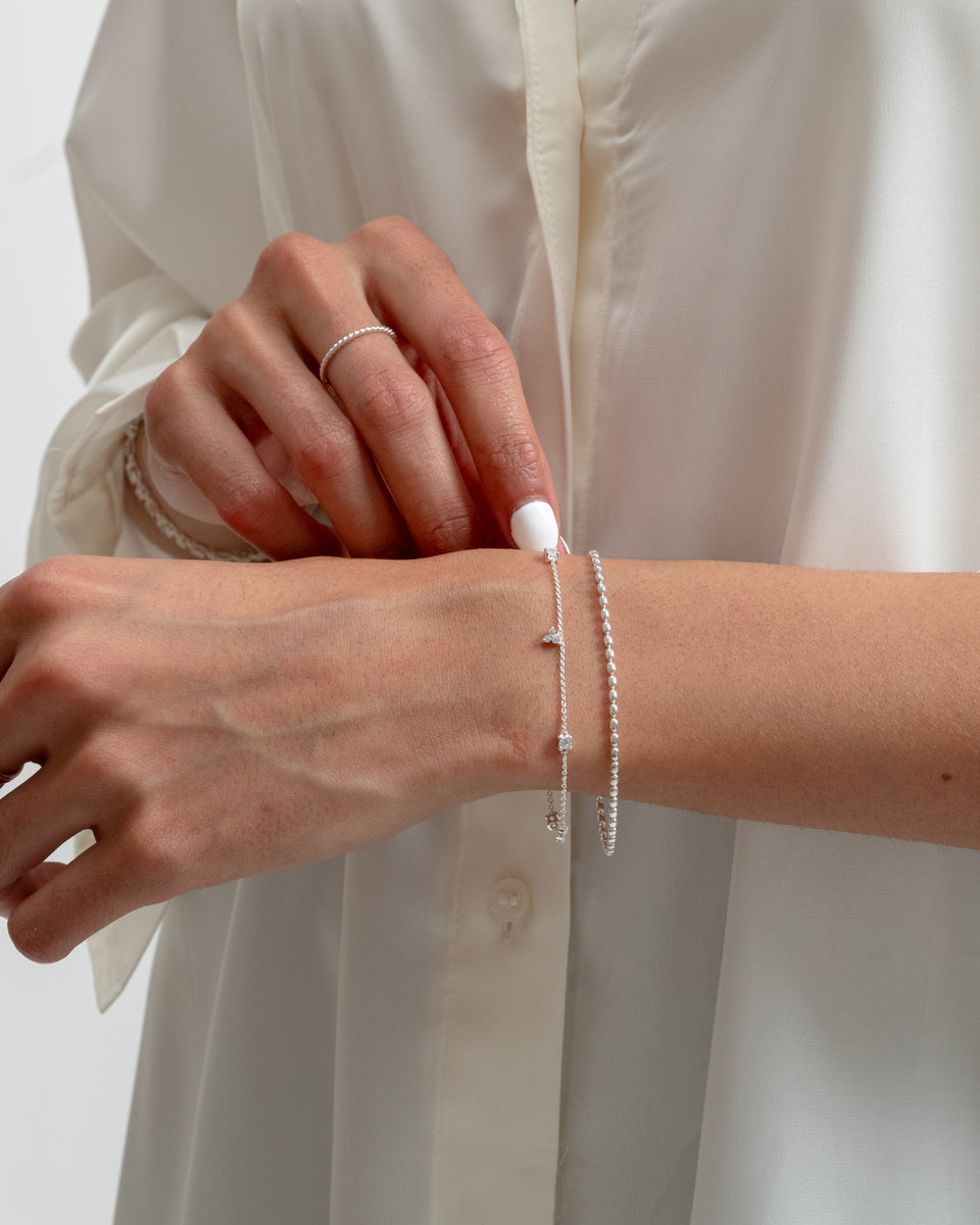 A close-up of a model wearing the Celestia Bracelet in silver, a delicate chain adorned with cubic zirconias. She has layered it with another silver beaded bracelet and is also wearing a thin, twisted silver ring.