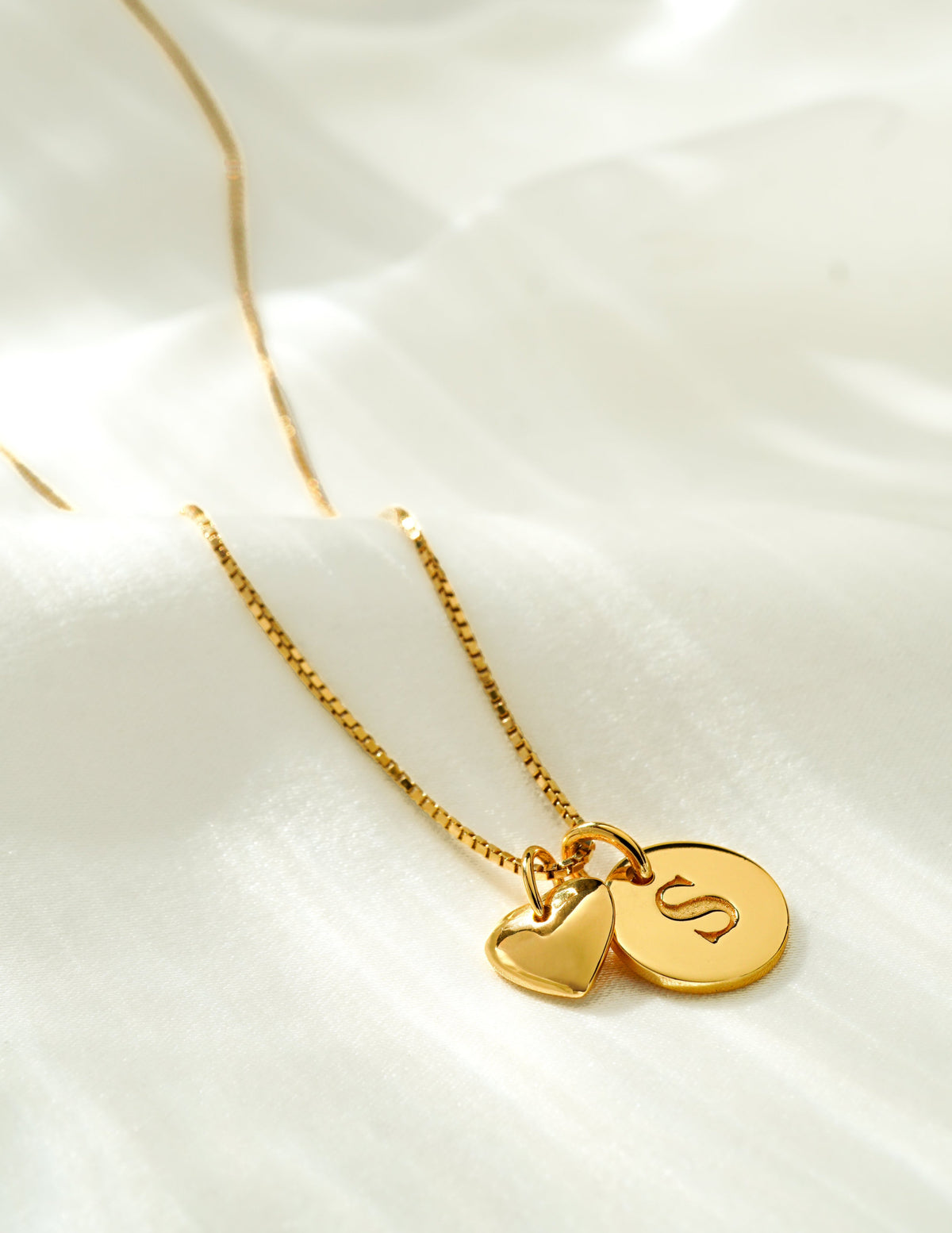 A product shot of a gold necklace on a medium box chain, styled with a Beloved Mini Letter 'S' pendant and a small heart pendant.