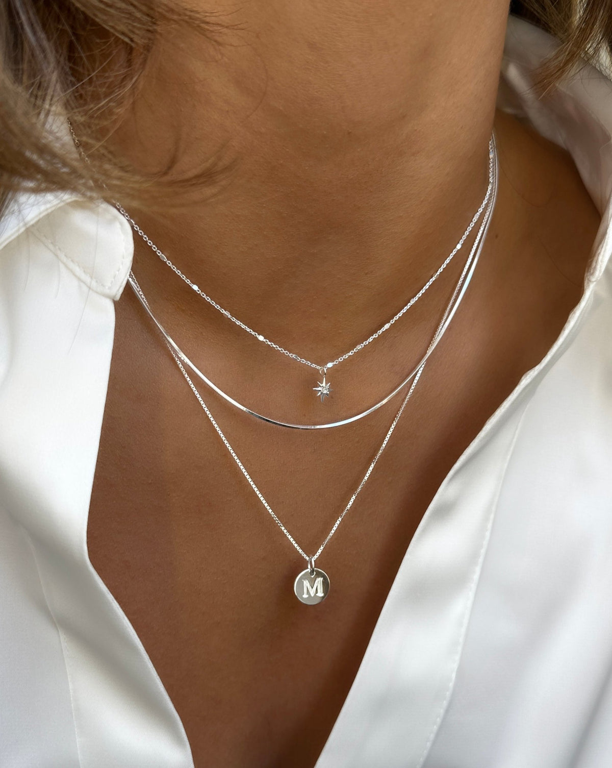 A close-up of a model wearing three layered silver necklaces: the North Star Necklace with a small star charm, the flat Sheer Mirror Necklace, and the Beloved Medium Box Chain with a round Beloved Mini Letter 'M' pendant.