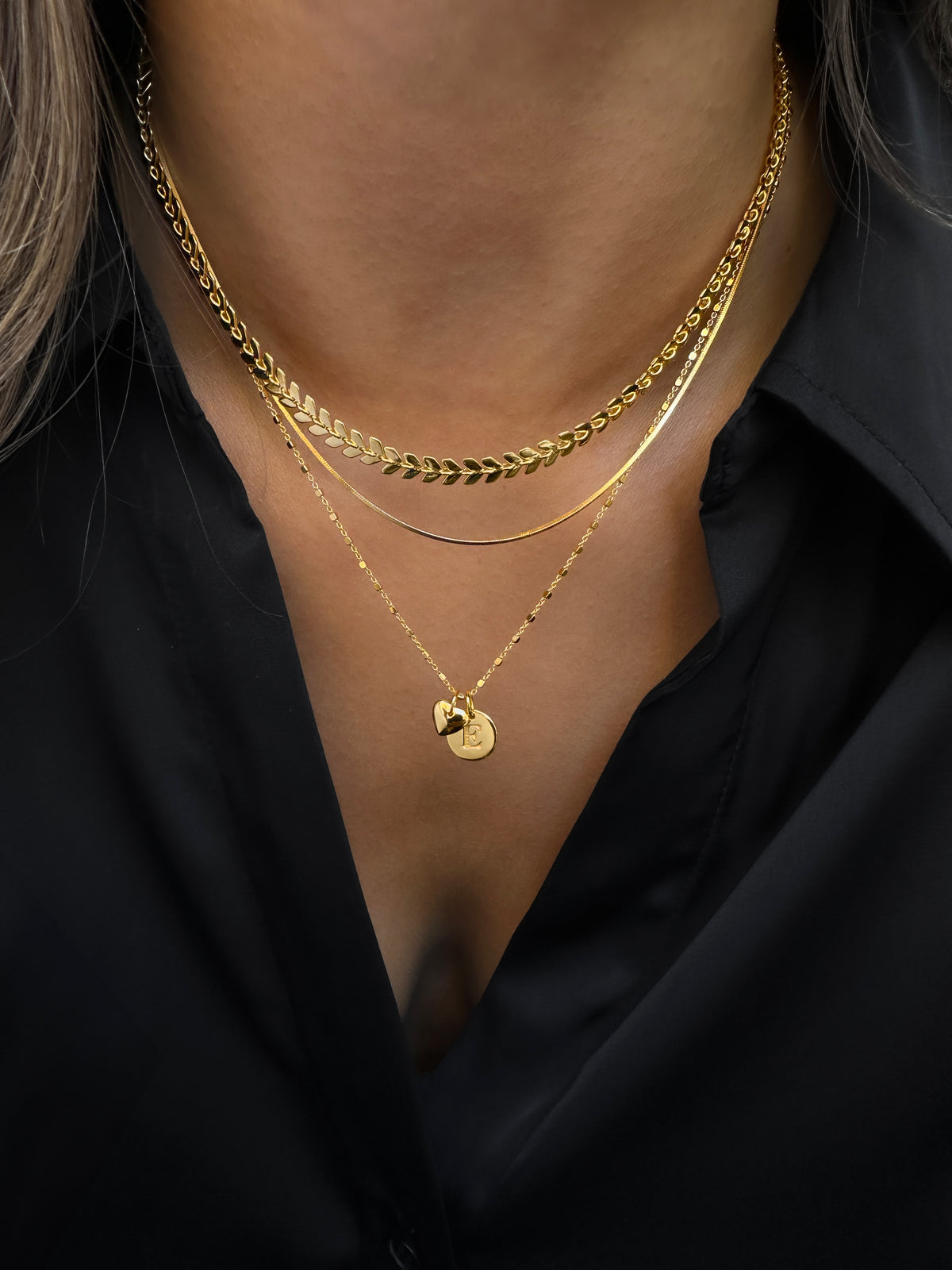 A close-up shot of a model wearing four layered gold necklaces, featuring a leaf-patterned chain, a flat snake chain, a simple delicate chain, and a longer chain with a round initial pendant and a heart charm.