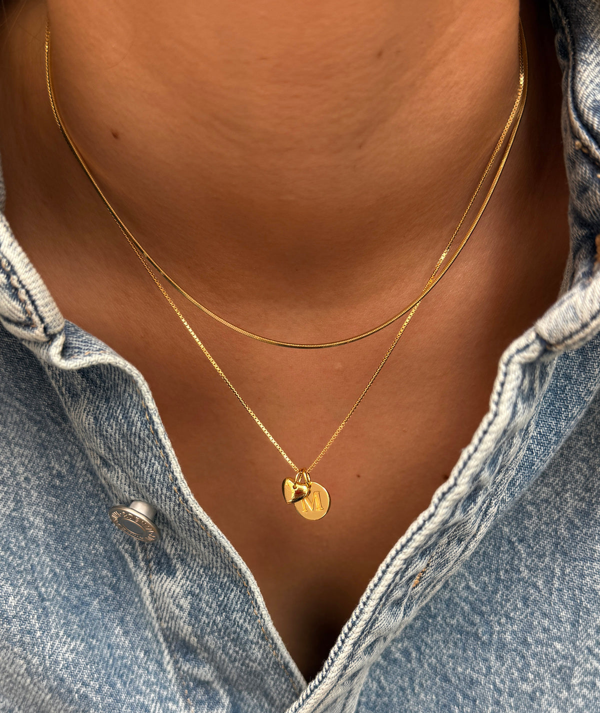 A close-up shot of a model wearing layered gold necklaces, including the Beloved Short Box Chain with a Beloved Mini Letter 'M' pendant and a gold Beloved Heart pendant.