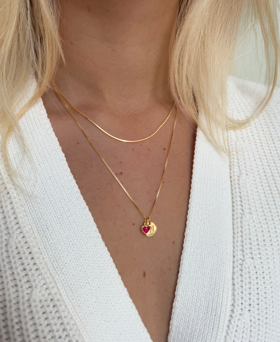 A close-up shot of a model wearing two layered gold necklaces: the Sheer Mirror Necklace and the Beloved Medium Box Chain personalized with a pink quartz heart pendant and a Beloved Mini Letter pendant.