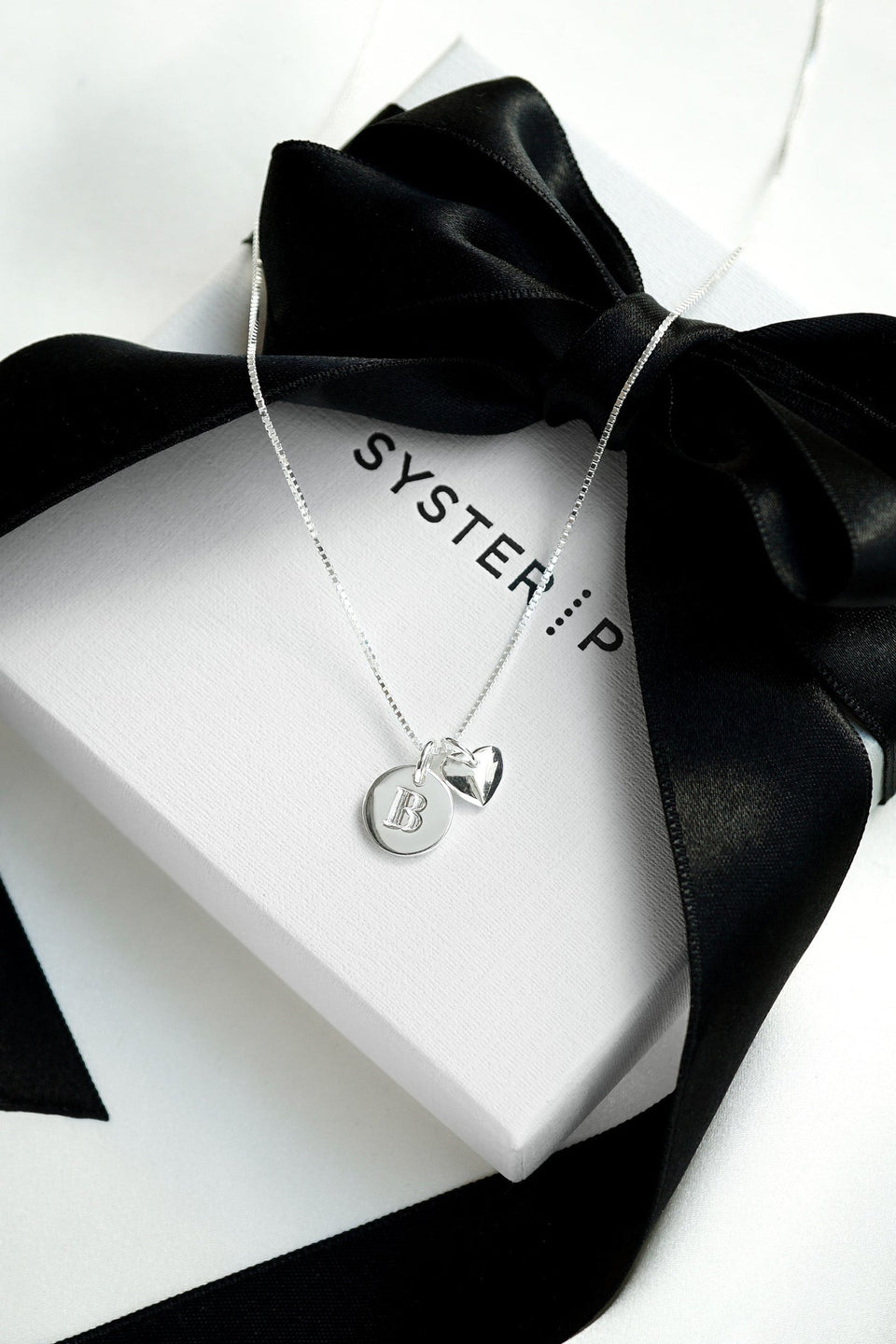 A product shot of a silver Syster P Beloved necklace with a medium box chain, featuring a mini letter 'B' pendant and a heart pendant, resting on a white gift box with a black ribbon.
