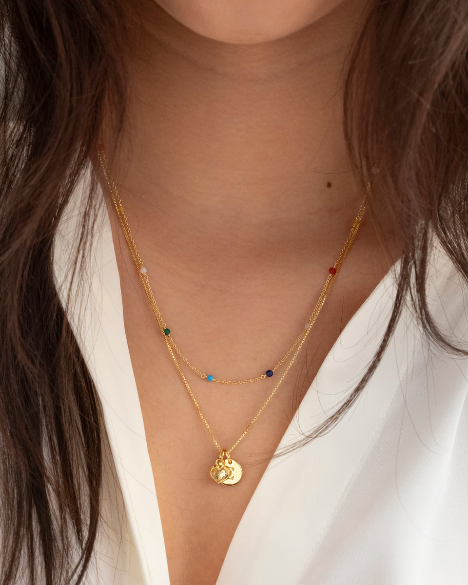 A close-up of a model wearing the Santa Monica Necklace in gold, featuring a fine chain with colourful gemstones, layered with a second gold necklace holding a crystal heart pendant and a disc charm with the letter E.