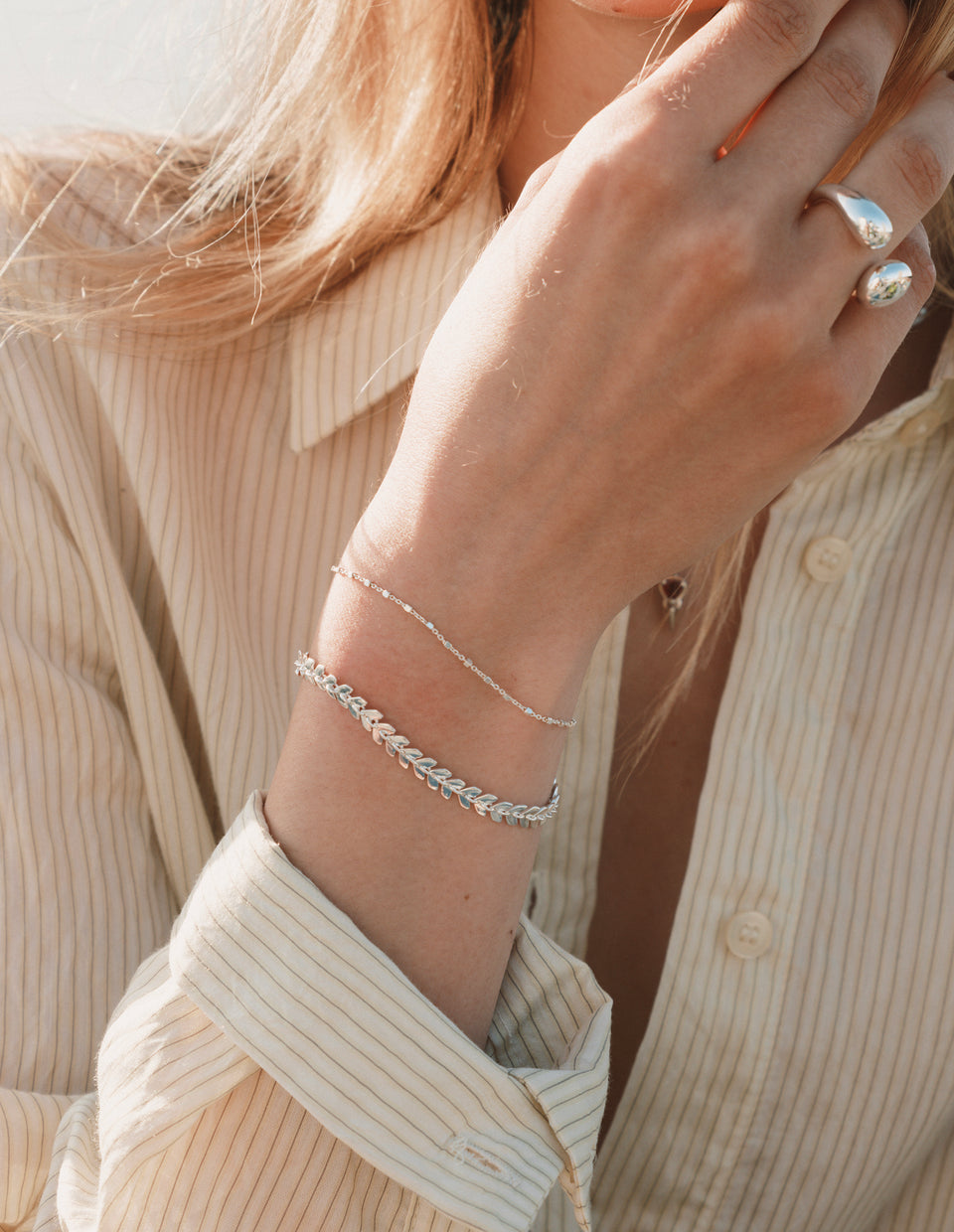 A close-up on a model's arm and hand, showing a stack of silver jewelry. She wears the leaf-patterned Layers Olivia Bracelet, the delicate Sheer Station Bracelet, and the Bolded Drops Ring.