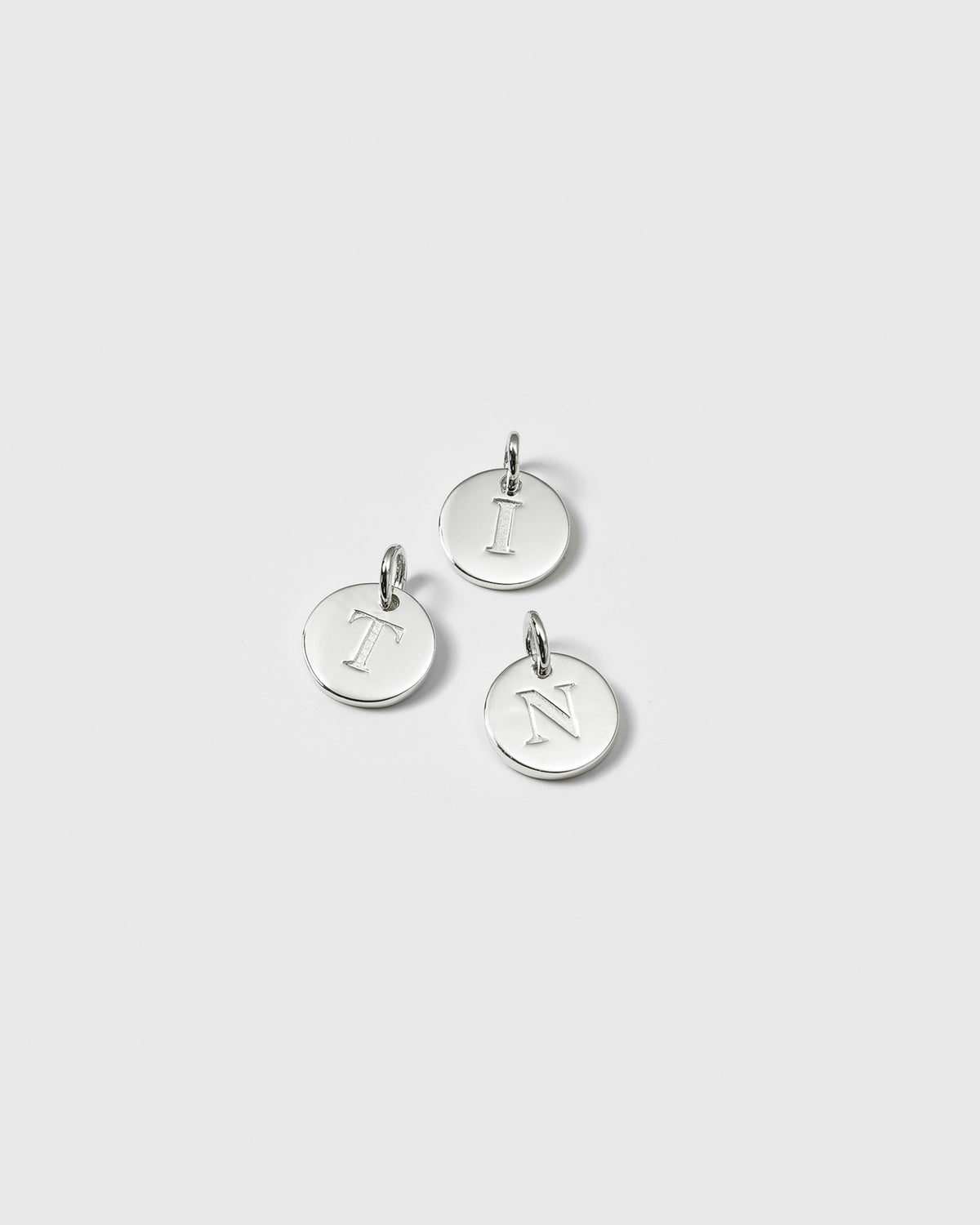 A product shot of the Beloved Mini Letter Silver pendants, showcasing three examples with the letters T, I, and N engraved on the small, circular charms.