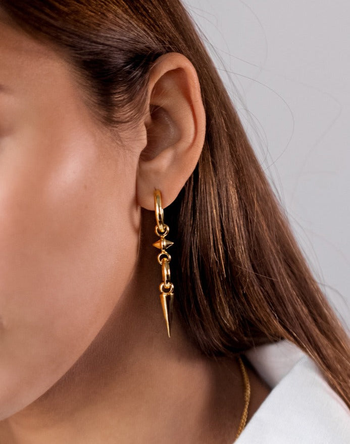 A close-up shot of a model wearing the Juno Long Single Earring in gold, which consists of a hoop with a long dangling pendant featuring several links and two prominent spikes. A delicate gold chain necklace is also visible.