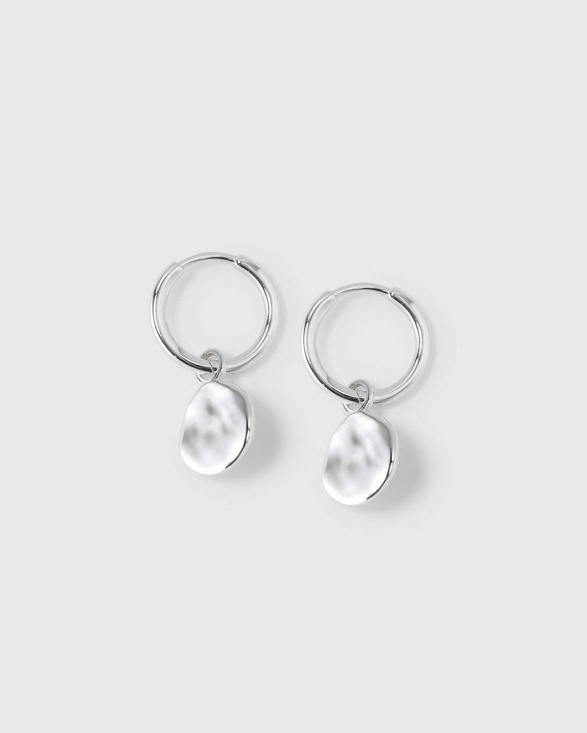 A product shot of the Minimalistica Hammered Earrings in silver, featuring a pair of small hoops, each with a dangling pendant with a hammered surface.