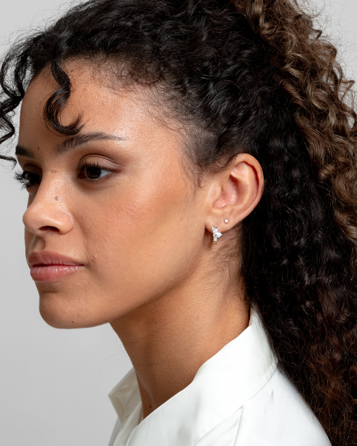 A close-up of a model wearing the Pam Hoops in silver, which are adorned with white topazes, and a small silver stud in her upper ear.