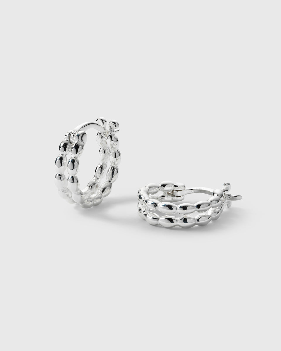 A product shot of the Lunetta Earrings in silver, with one earring standing up and the other lying down to showcase their unique design of two parallel bands connected by a pattern of small ovals.