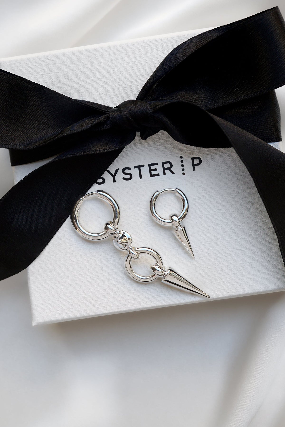 A product shot of the Juno Long Earring and the Juno Single Hoop Earring in silver, displayed on a white Syster P gift box to highlight their edgy design with dangling spikes.