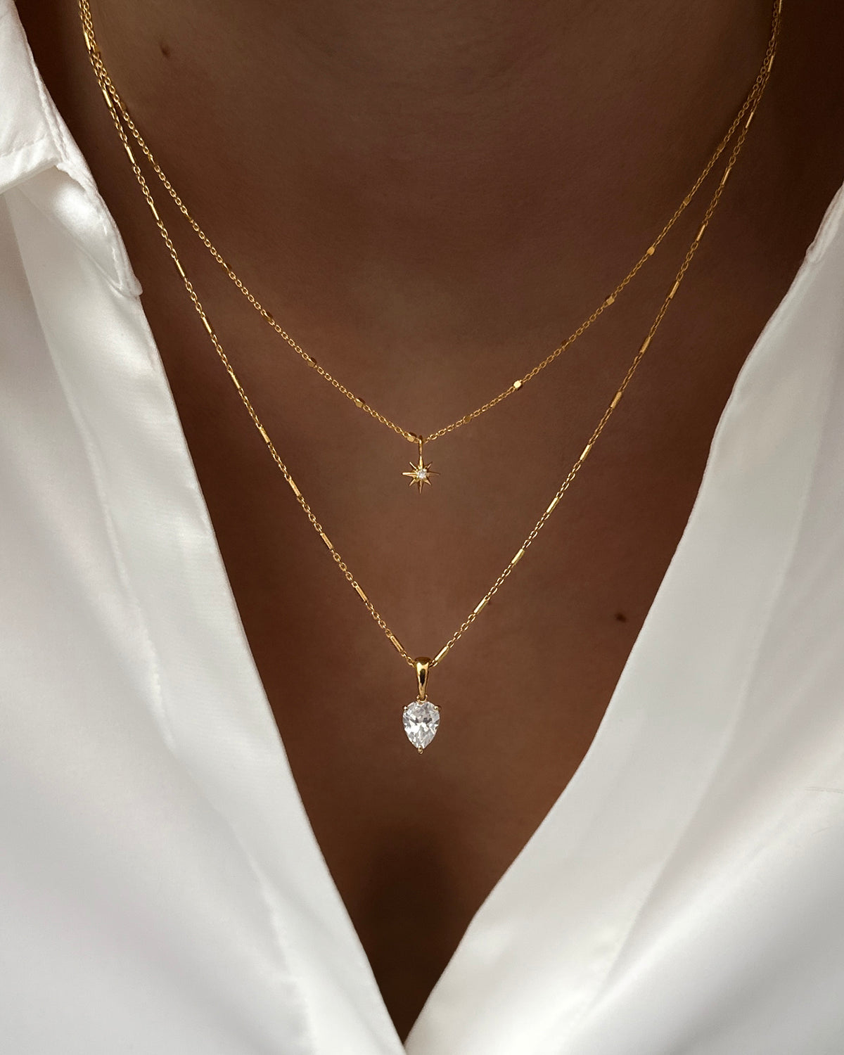 A close-up of a model wearing two layered gold necklaces against her skin and a white shirt; the shorter North Star Necklace with a star pendant and the longer Katie Necklace with a teardrop crystal.