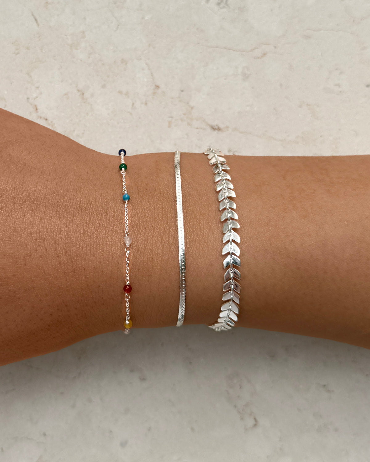 A close-up of a model's wrist styled with three layered silver bracelets from Syster P: a thin chain with colorful beads, a smooth Herringbone Bracelet, and one with a leaf-like pattern.