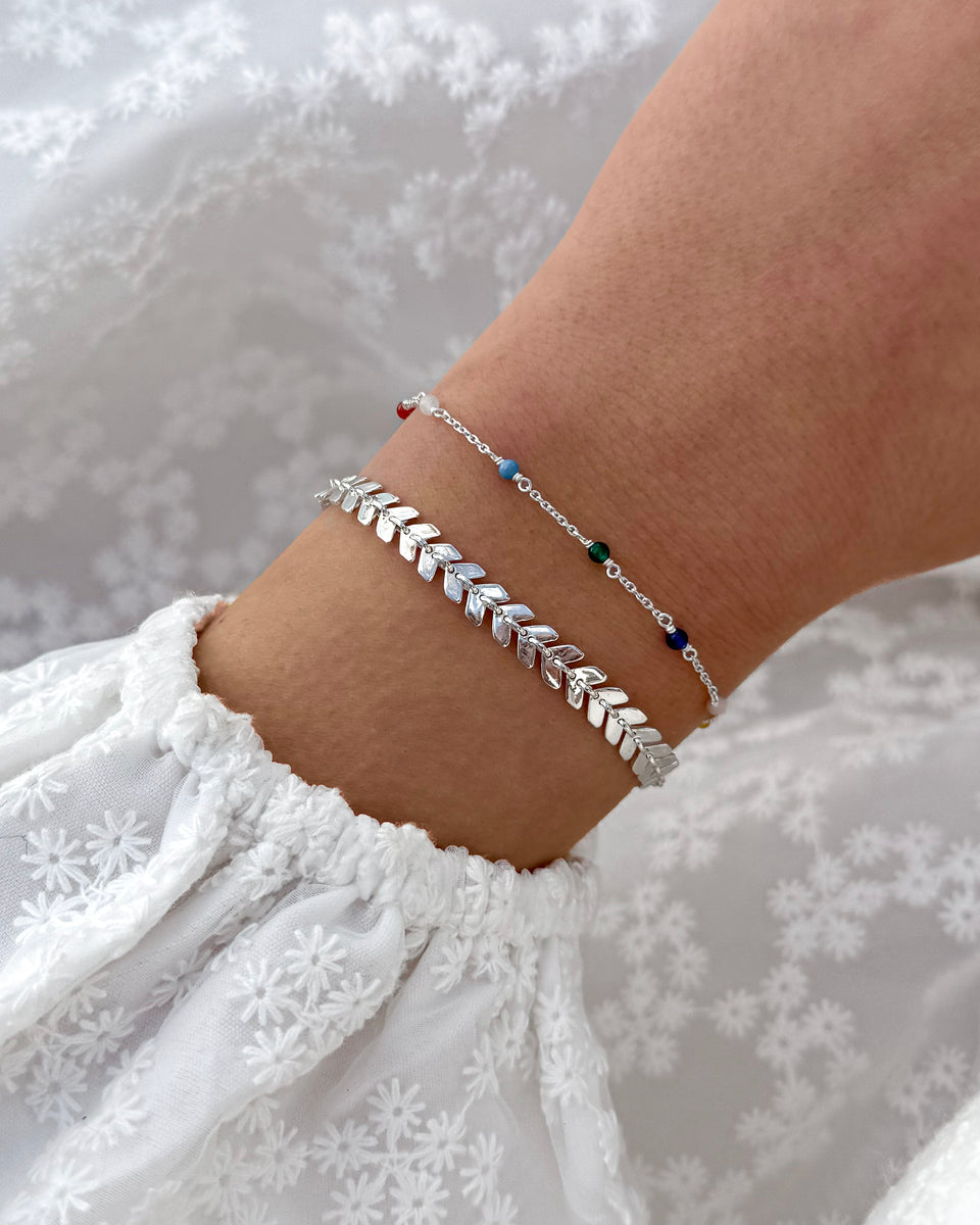 A close-up of a model's wrist wearing the Layers Olivia Bracelet in silver, featuring a pattern of small leaf-like links. It is layered with the Santa Monica Bracelet, a delicate silver chain with small, colorful beads.