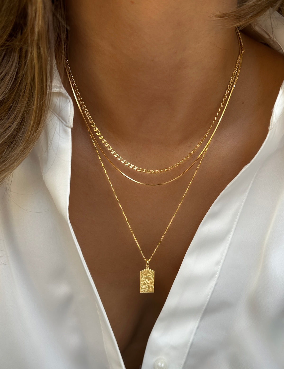 A close-up of a model wearing a layered set of three gold necklaces: the Lucky Coin Keep Dreaming Necklace with its rectangular pendant, the flat Sheer Mirror Necklace, and the Sheer Love curb chain necklace.