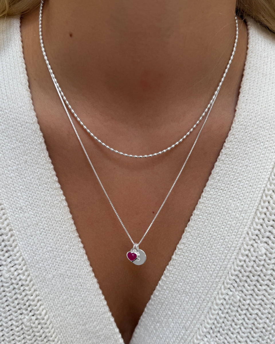 Silver layered necklaces: a delicate beaded chain and a box chain with a pink quartz heart and letter pendant.