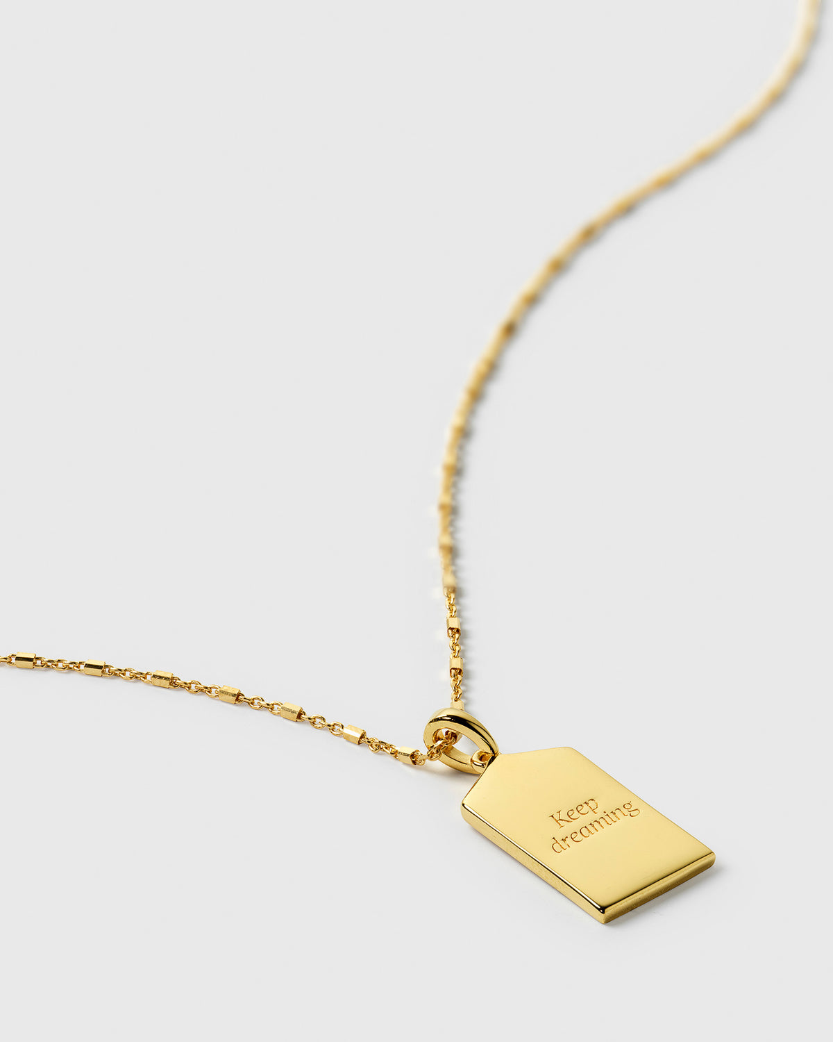 A close-up shot of the Lucky Coin Keep Dreaming Necklace in gold, showing the back of the rectangular tag pendant engraved with the words "Keep dreaming".