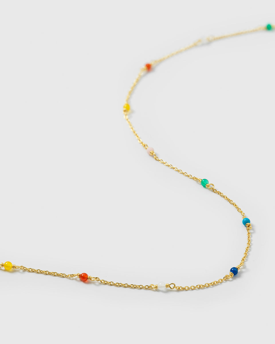 A close-up product shot of the Santa Monica Necklace Gold, laid out to showcase its delicate gold chain adorned with small, vibrant, multi-colored gemstone beads: Yellow Jade, Pink Opal, Sodalite, Green Onyx, Blue Color Reconstituted Turquoise, White Agate, and Carnelian.