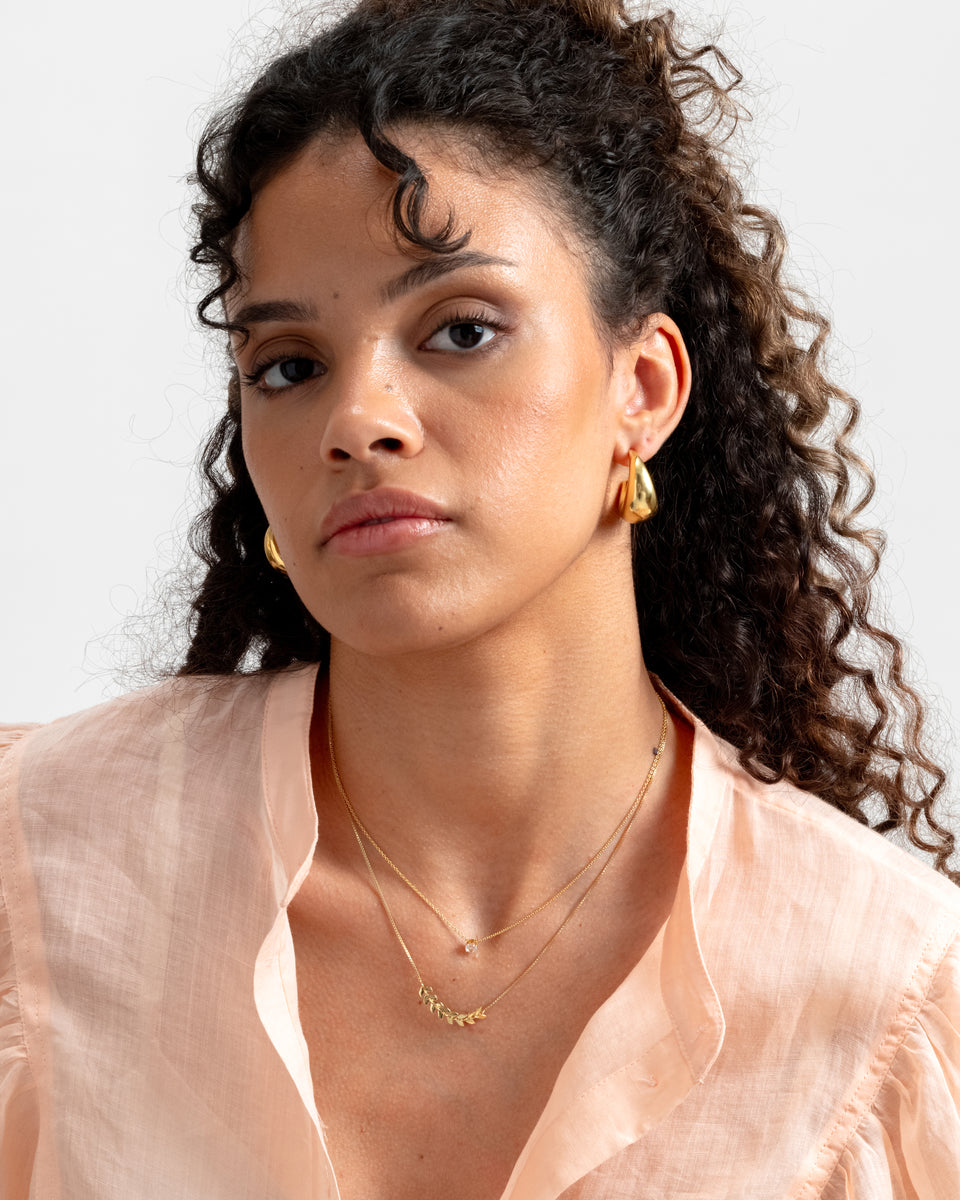 A model wearing the gold Minimalistica Solo Nova Necklace, featuring a delicate chain and a single white topaz, layered with another gold necklace with a leaf design and paired with chunky gold hoop earrings.