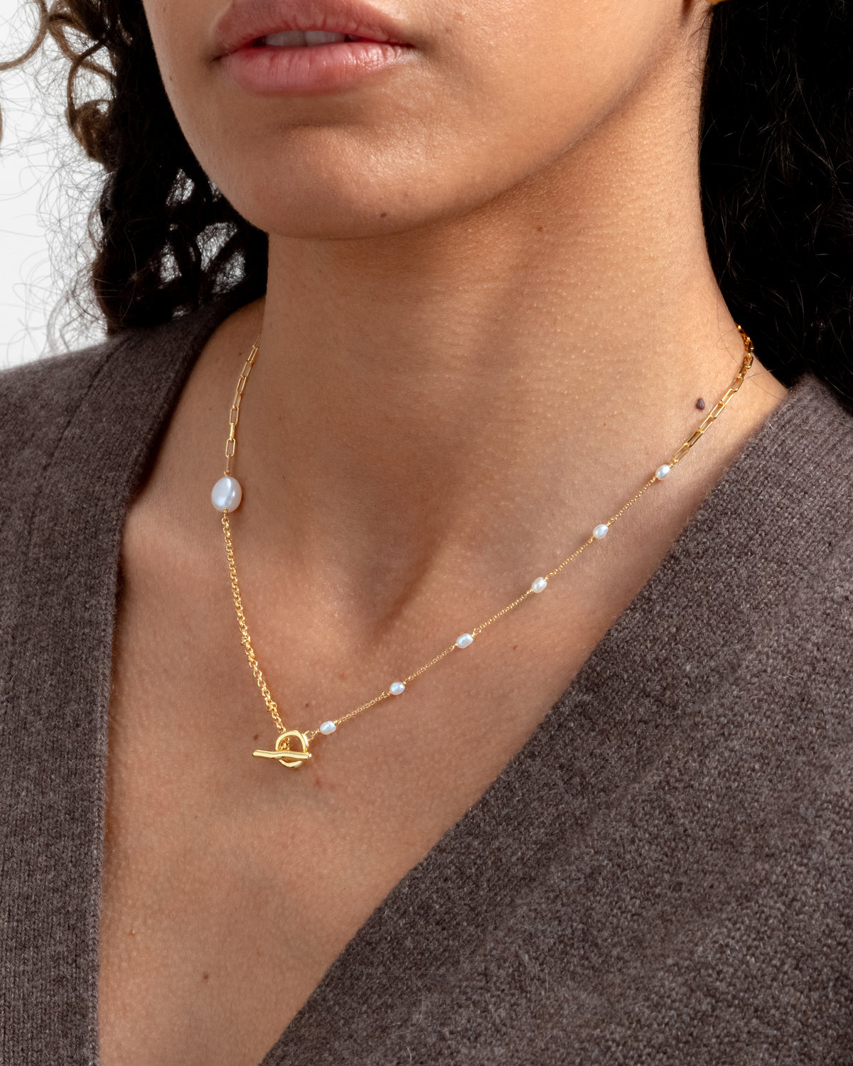 Close-up on a model's neck, wearing the Paxton Short Necklace Gold. The necklace features a mix of chain styles, baroque freshwater pearls, and a toggle clasp.