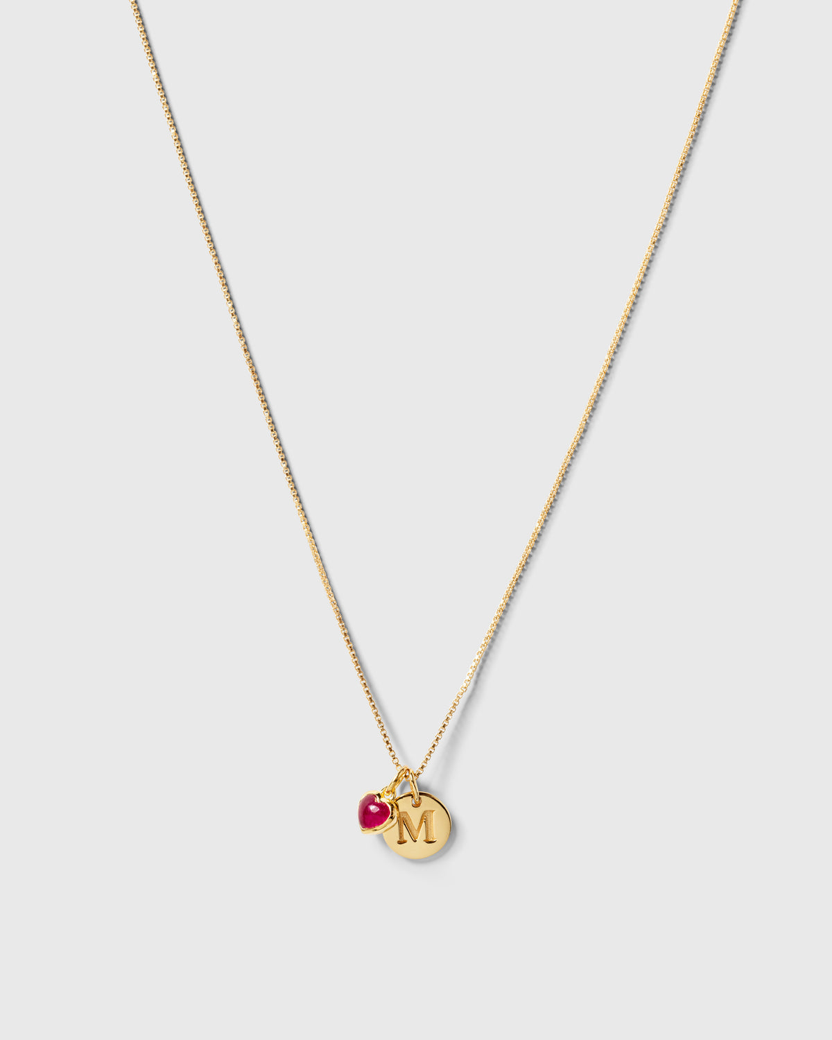 A product shot of a gold necklace featuring the Beloved Heart Pendant with a pink quartz stone, styled alongside a round gold pendant with the initial 'M'.