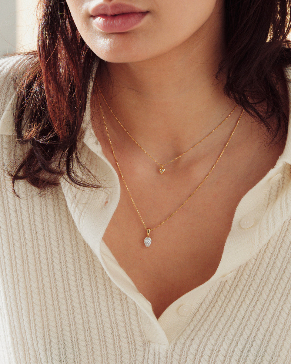 A model shot of the Katie Necklace in gold, featuring a drop-shaped cubic zirconia pendant, layered with a shorter, delicate gold necklace with a small heart pendant.