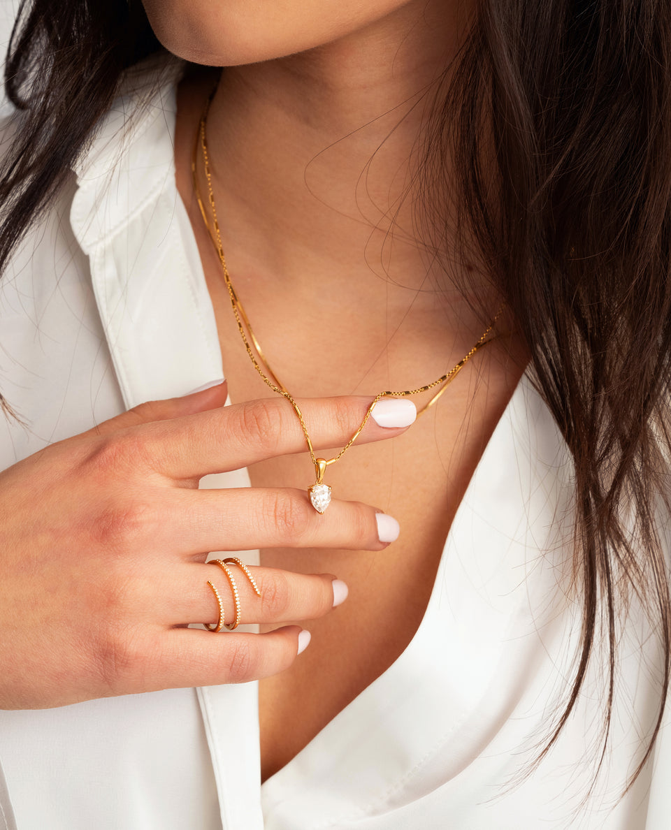 A model wears the Katie Necklace Gold, featuring a drop-shaped cubic zirconia pendant on a unique chain, styled with a sparkling gold wrap ring.