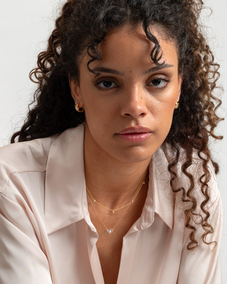 A close-up of a model wearing the Celestia Necklace in gold, a delicate chain with small cubic zirconias, layered with a second gold necklace and paired with matching gold hoop earrings and a stud.