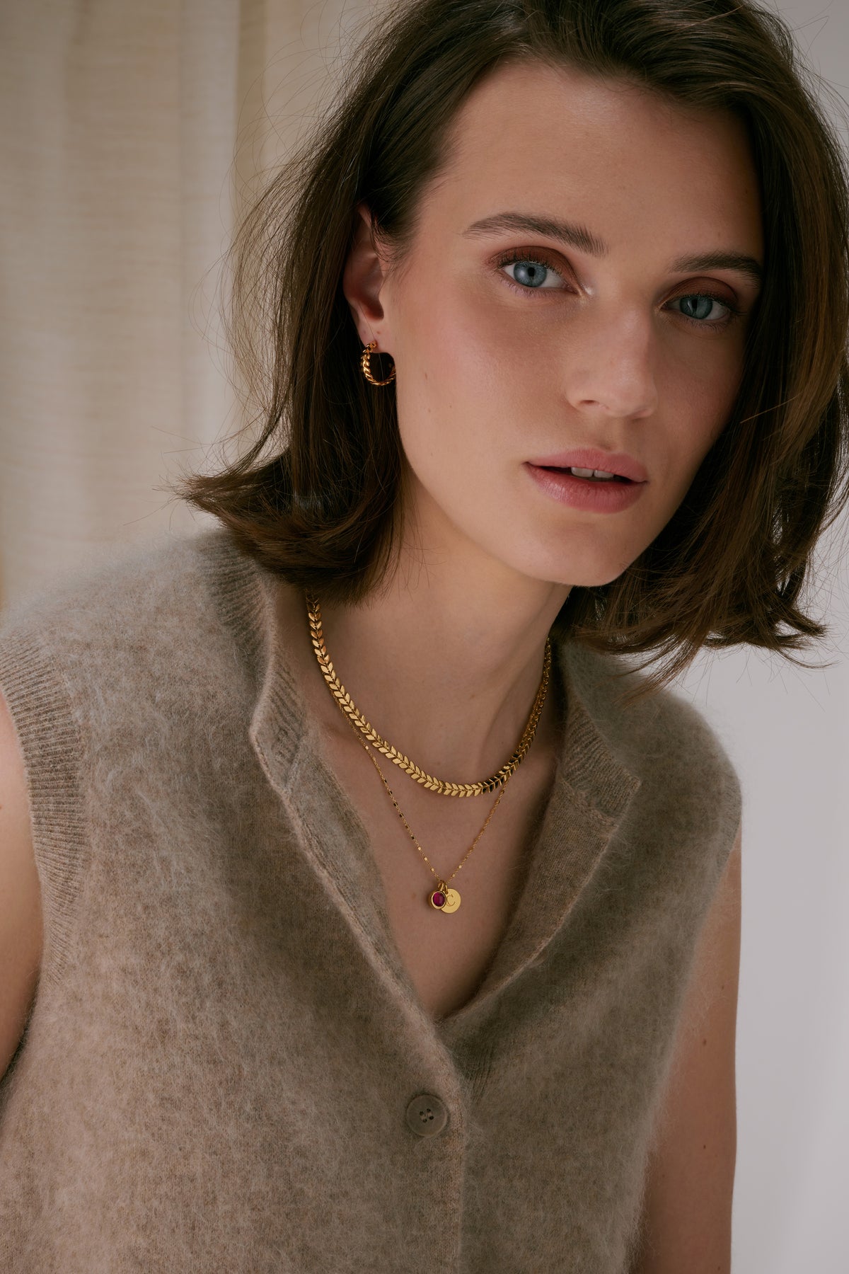 The gold Beloved Birthstone July pendant with a red ruby, styled with a layered gold leaf chain necklace, a gold initial pendant, and small gold twisted hoop earrings.
