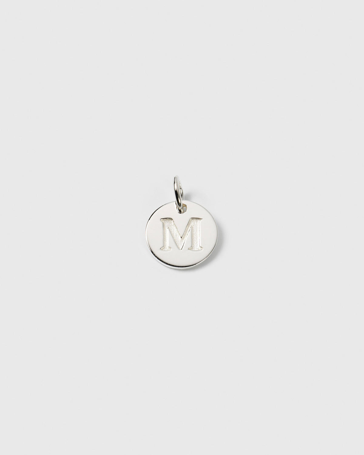 A product shot of the Beloved Mini Letter M charm in silver, a small, circular pendant with a capital M engraved on its surface.