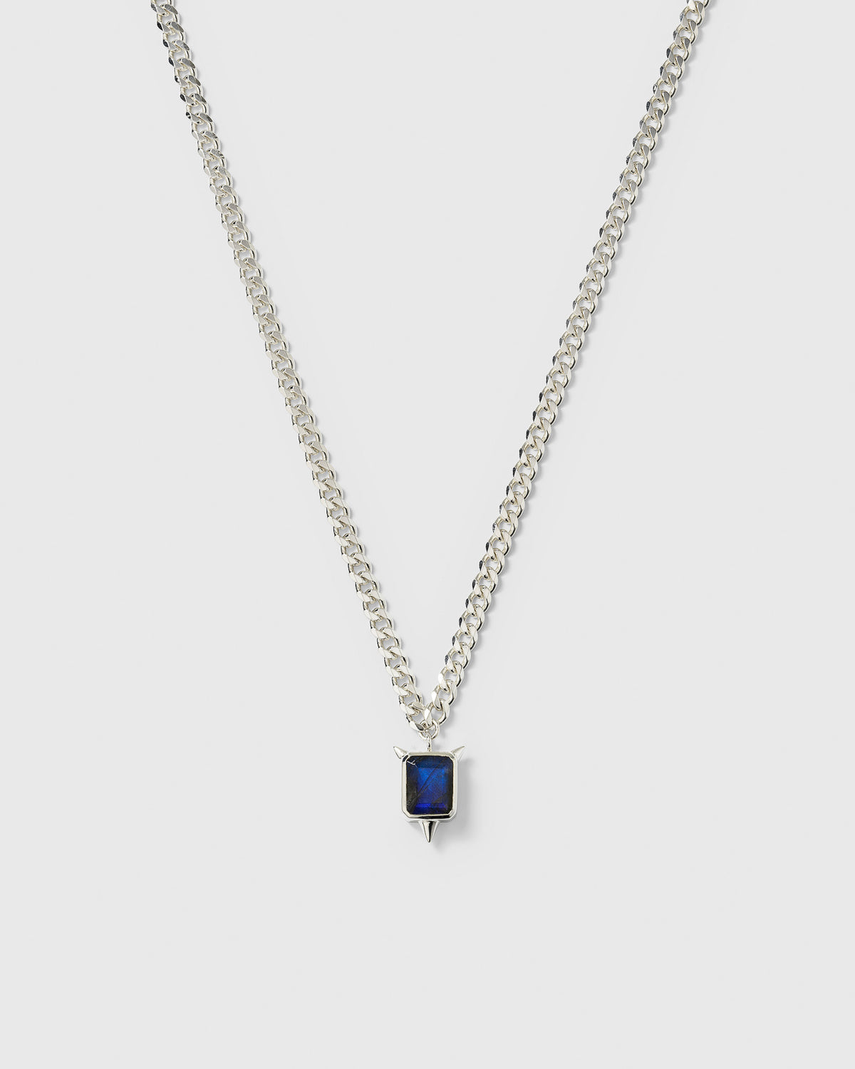 A product shot of the Drama Necklace in silver, featuring a heavy chain and a pendant with a rectangular labradorite stone and three edgy spikes.