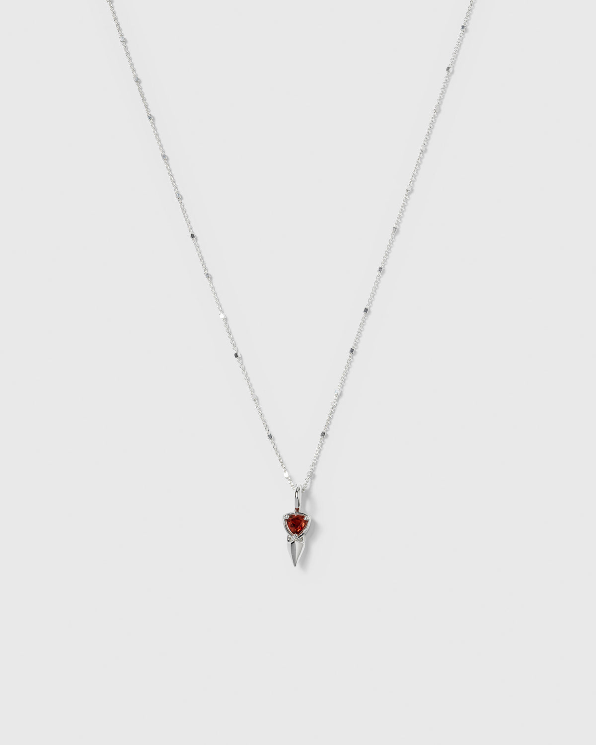 A product shot of the Mrs H Spike Necklace in silver, displaying its fine, beaded chain and a pendant composed of a sharp spike and a vibrant red garnet.