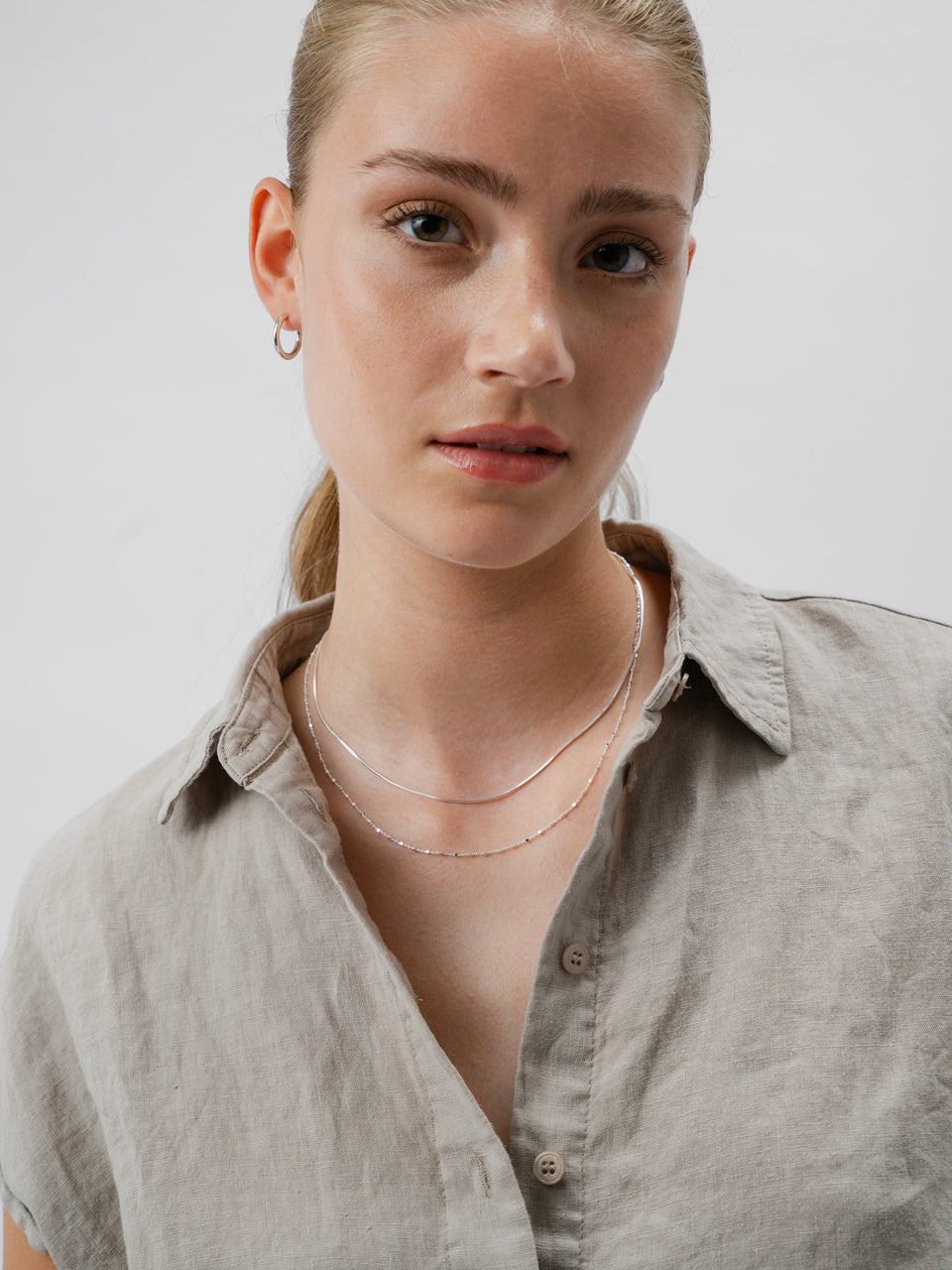 A necklace in silver, a smooth chain, layered with another delicate silver necklace and small silver hoop earrings.