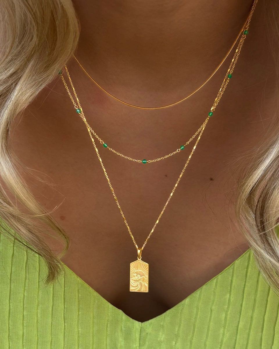 A model wearing a stack of three layered gold necklaces: the Santa Monica necklace with green onyx stones, the Sheer Mirror chain, and the Keep Dreaming necklace with a rectangular pendant.
