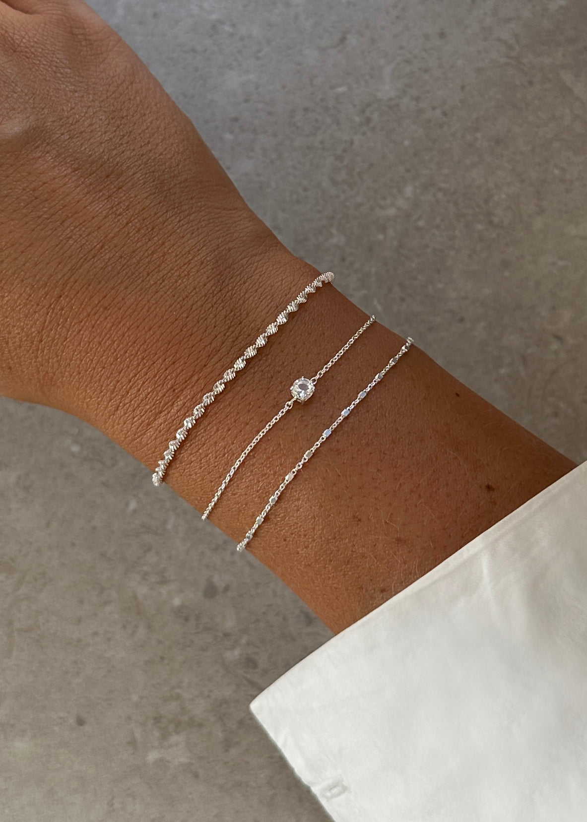 A close-up of a model's wrist wearing a stack of three silver bracelets: the Herringbone Twisted Bracelet, the Minimalistica Solo Nova Bracelet with a single stone, and the delicate Sheer Station Bracelet.