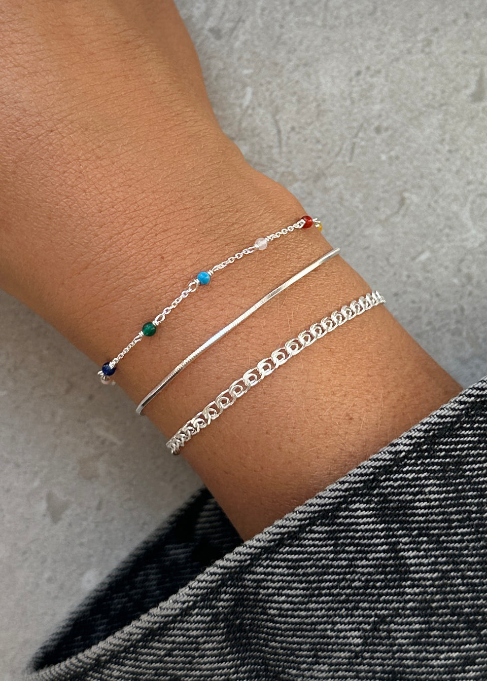 A model's wrist styled with a stack of three Syster P silver bracelets: the Santa Monica Bracelet with colorful gemstones, the simple Sheer Mirror chain bracelet, and the Sheer Love linked chain bracelet.