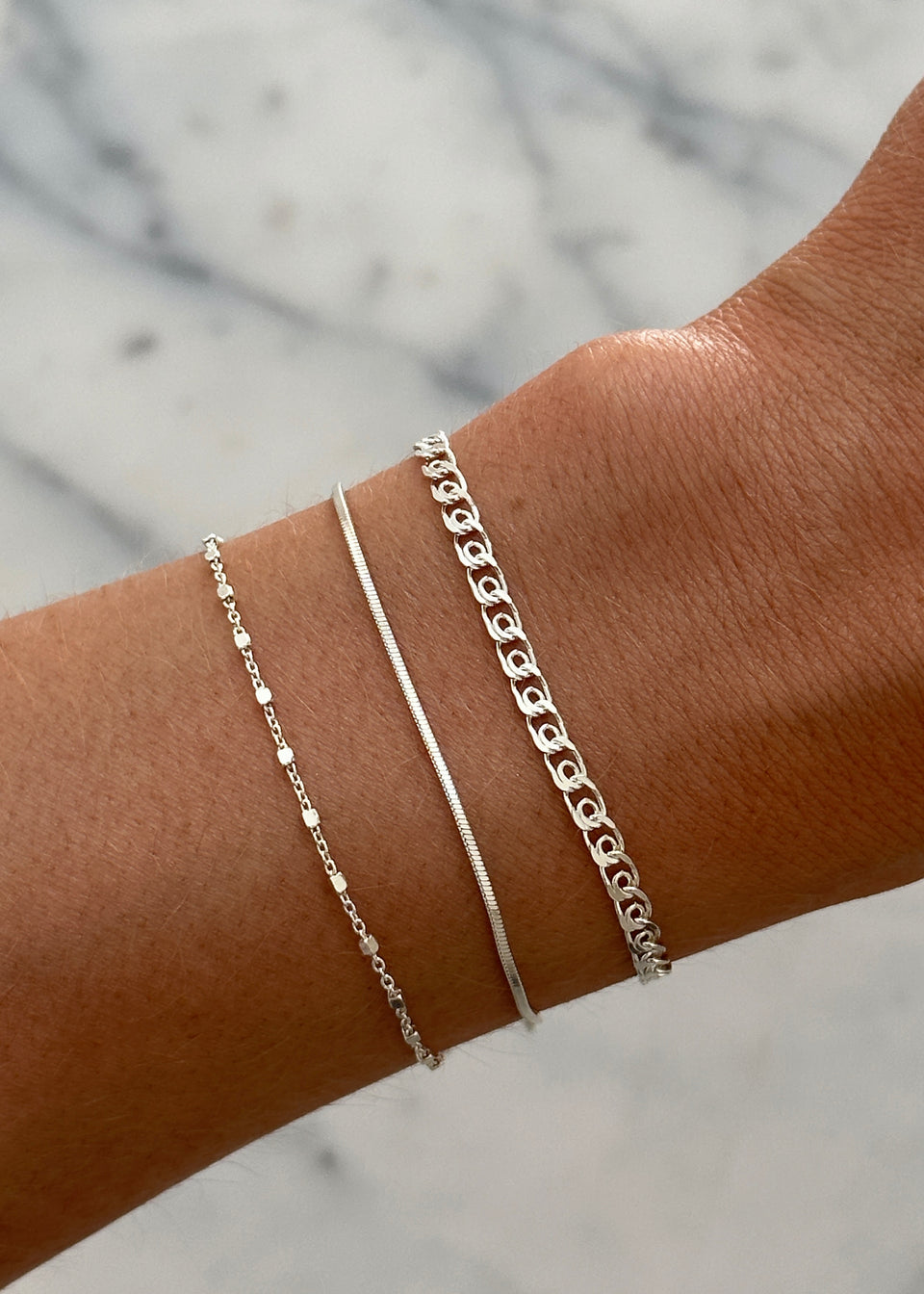 A close-up of a model's wrist wearing a stack of three silver bracelets: the Sheer Love Bracelet, the Sheer Mirror Bracelet, and the Sheer Station Bracelet.