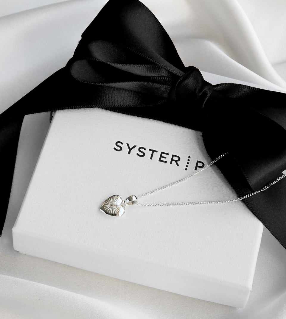 A product shot of the Sunburst Heart Necklace in silver resting on a white Syster P jewelry box tied with a black ribbon, showcasing its heart-shaped pendant with a sunburst pattern.