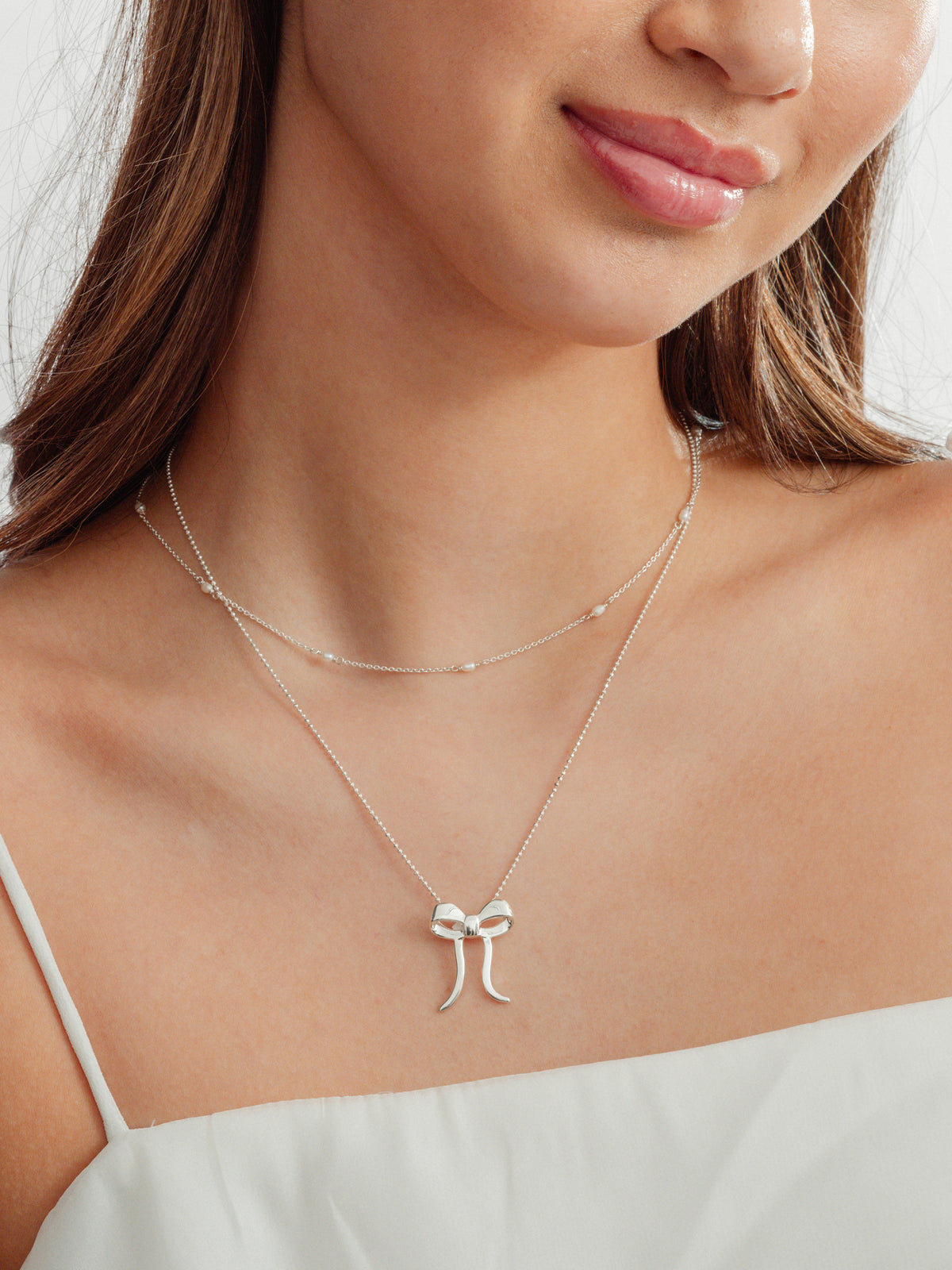 A close-up shot of a model wearing the Treasure Multi Pearl Necklace layered with the Rosie Necklace, both in silver. The Treasure necklace features small freshwater pearls on a delicate chain, while the Rosie Necklace has a bow pendant.