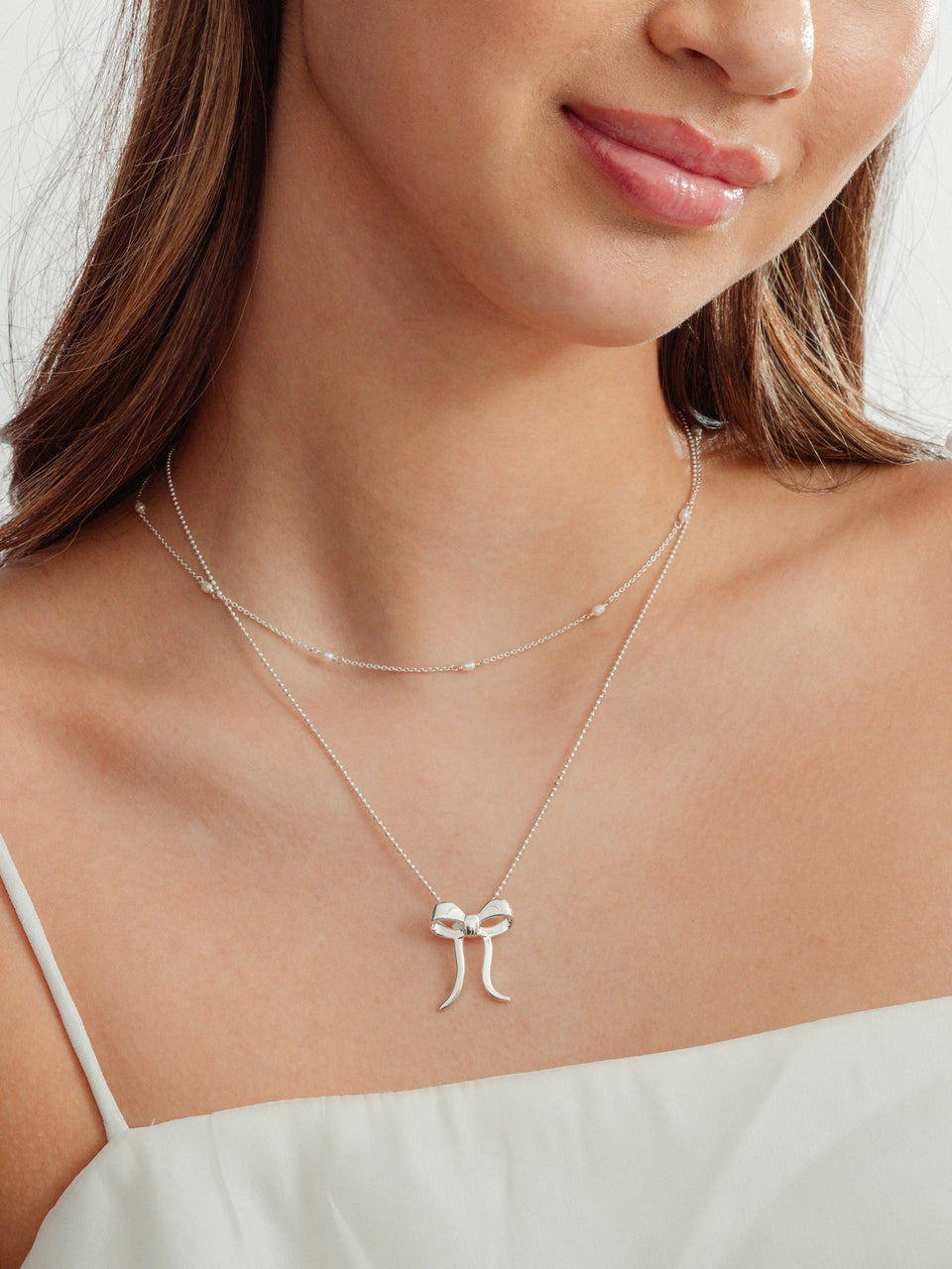 A close-up shot of a model wearing the Treasure Multi Pearl Necklace layered with the Rosie Necklace, both in silver. The Treasure necklace features small freshwater pearls on a delicate chain, while the Rosie Necklace has a bow pendant.