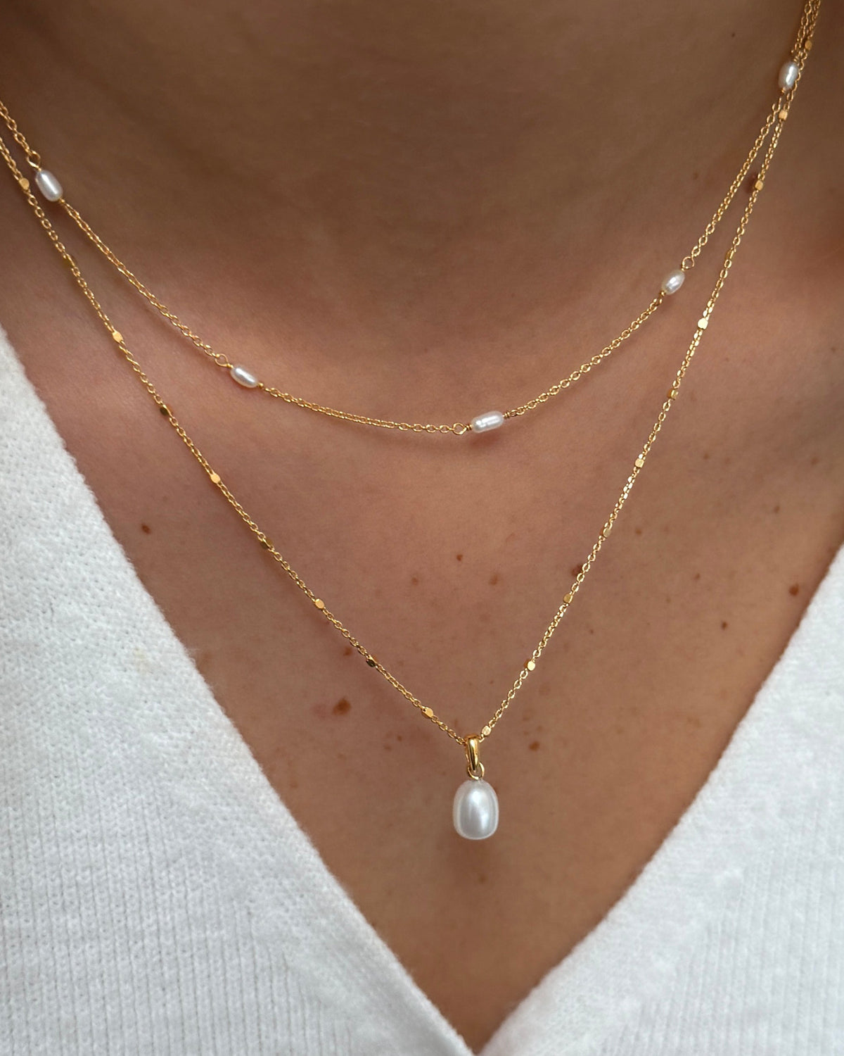 A close-up of a model wearing two layered gold pearl necklaces from the Treasure collection. The shorter necklace features two fine chains with small, interspersed pearls, while the longer necklace has a single teardrop pearl pendant.