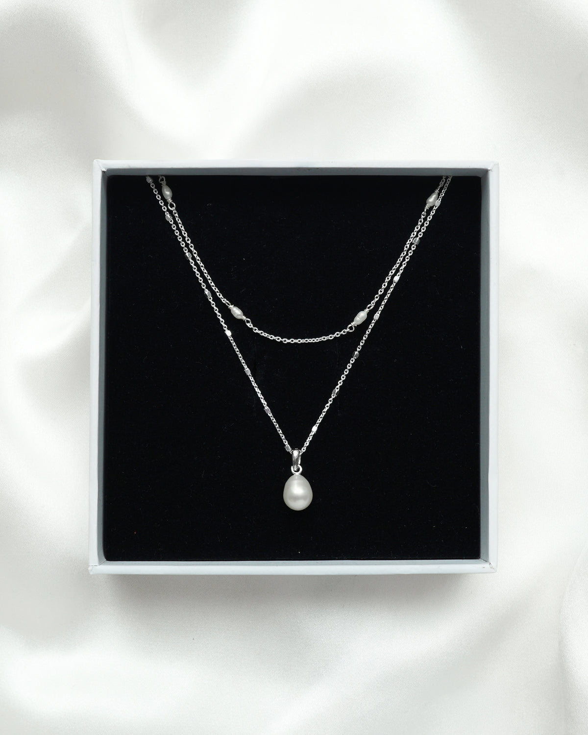 The Treasure Necklace Set Silver is displayed in its white Syster P gift box, showcasing the two layered necklaces: one with a single teardrop freshwater pearl and another with multiple small pearls along its silver chain.