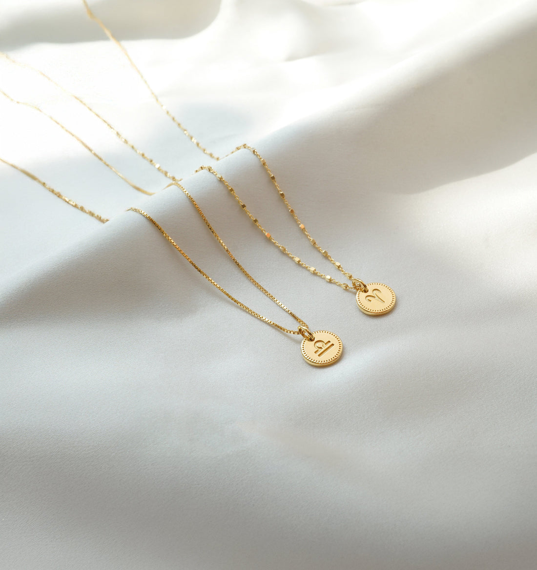 Two gold necklaces with zodiac pendants placed on silk