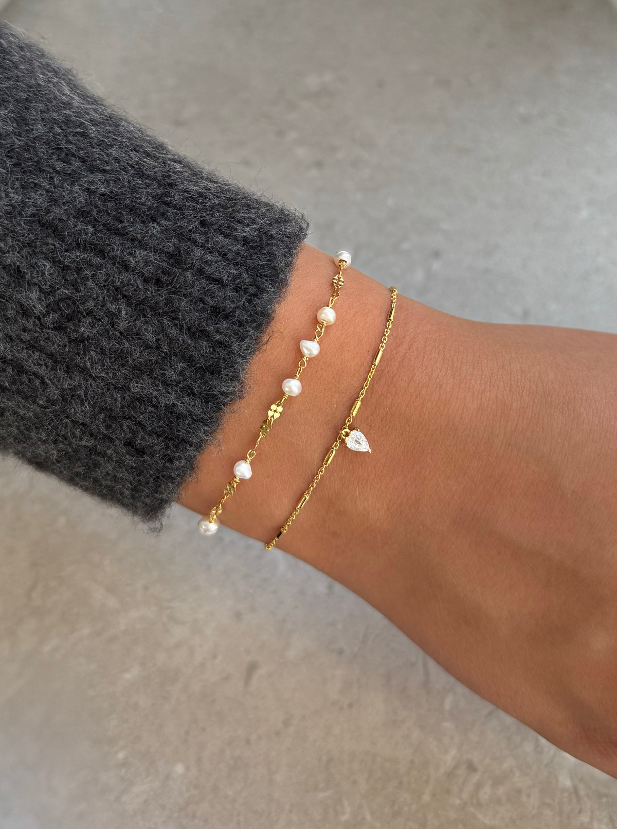 A close-up shot of a model's wrist wearing the gold Aurelia Bracelet, featuring a unique chain with small freshwater pearls, stacked with the gold Katie Bracelet, a fine chain with a teardrop-shaped crystal charm.