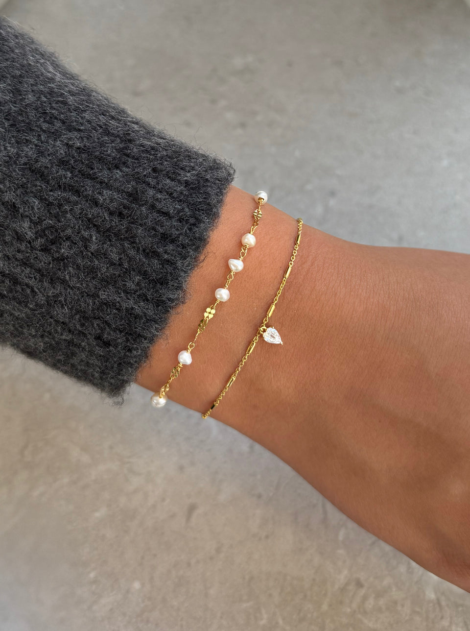 A close-up shot of a model's wrist wearing the gold Aurelia Bracelet, featuring a unique chain with small freshwater pearls, stacked with the gold Katie Bracelet, a fine chain with a teardrop-shaped crystal charm.